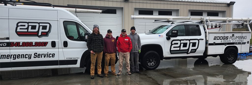 Four men stand in front of two 2 Dogs Plumbing service vehicles parked outside a garage on a wet pavement.